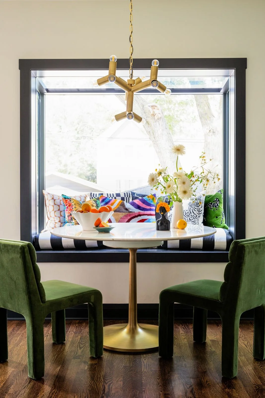 A cozy dining nook with a white round table, two green velvet chairs, and a window seat with colorful pillows and a striped cushion. A flower vase with white flowers and small decorative items are on the table, and a modern gold chandelier hangs abov