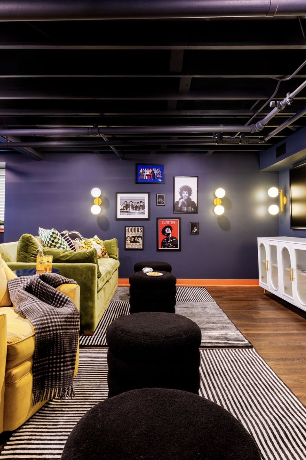 A cozy living room with a dark blue accent wall decorated with framed photos and posters, a mustard yellow sofa with patterned pillows, black ottomans, and a white cabinet, illuminated by wall-mounted lights and a ceiling glow, with striped rugs and 