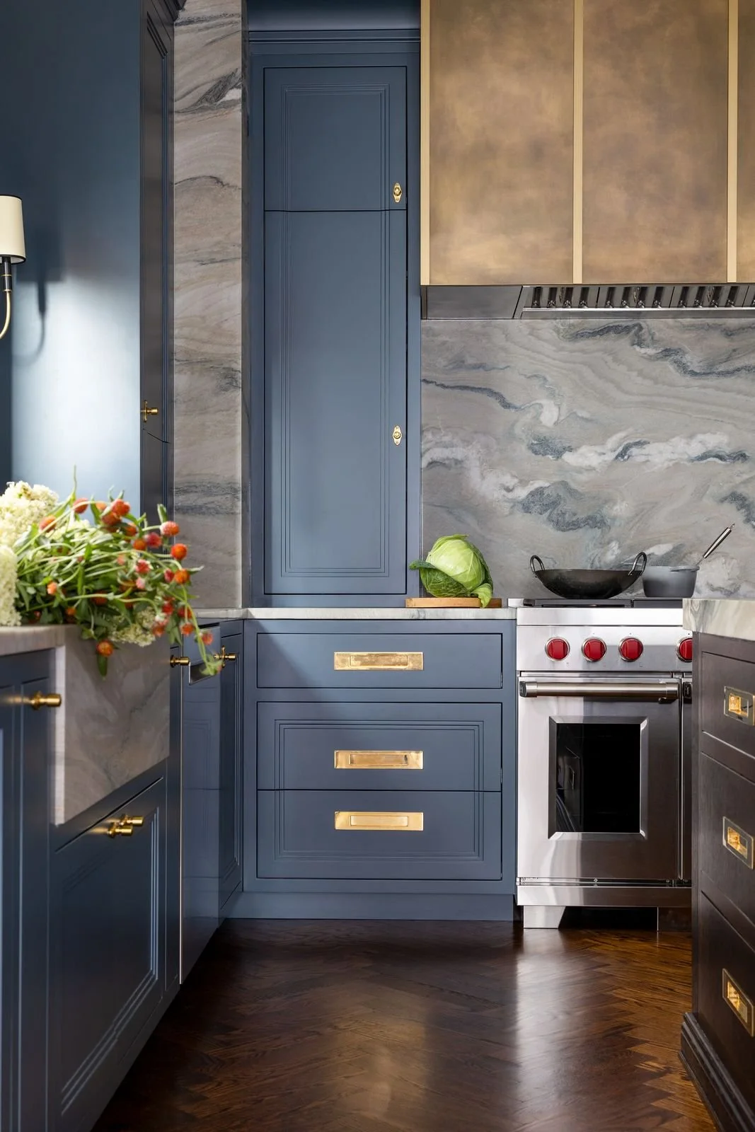 A kitchen with blue cabinets, gold handles, a marble backsplash, a stainless steel oven with red knobs, a cabbage on a cutting board, a black wok with a spoon, and floral arrangement on the counter.