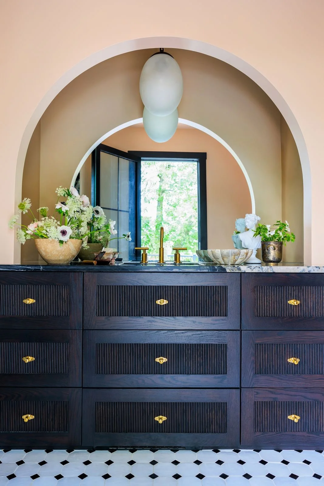A bathroom vanity with dark wood drawers, gold knobs, and a black and white tiled floor. The vanity has a marble countertop with a gold faucet, decorative bowls, a vase with white and purple flowers, and a small statue. Behind the vanity, there are t