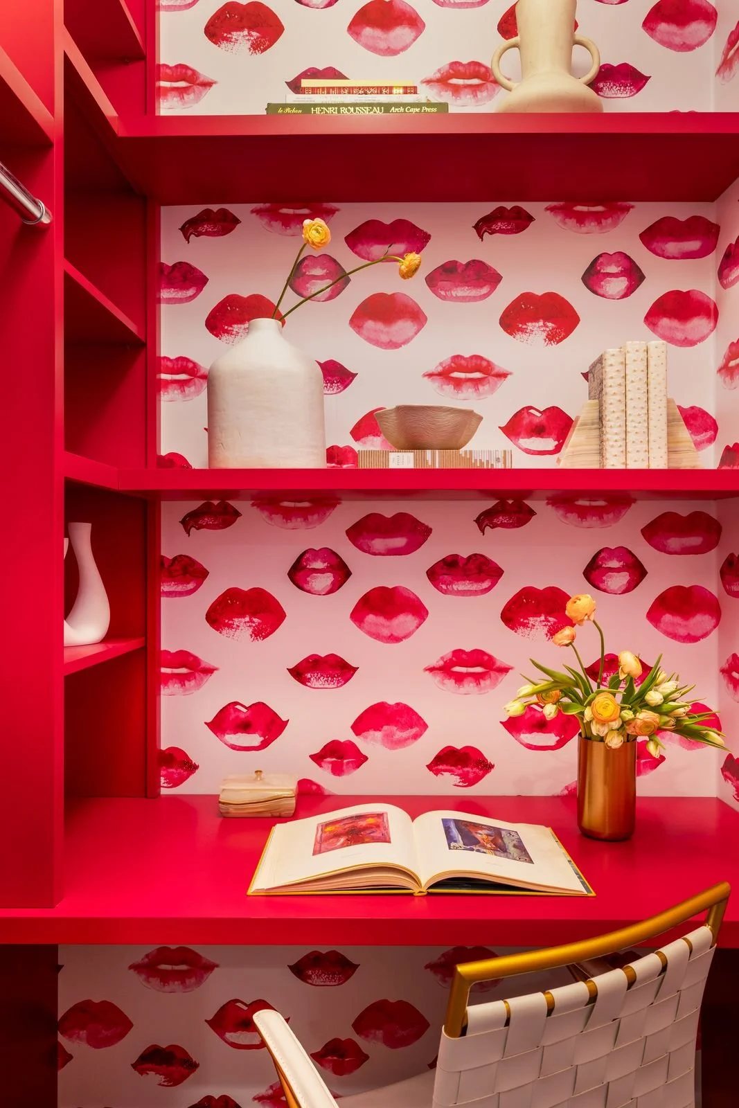 Pink wall with lipstick kiss pattern, red desk with open book, gold vase with tulips, and decorative objects on red shelves.