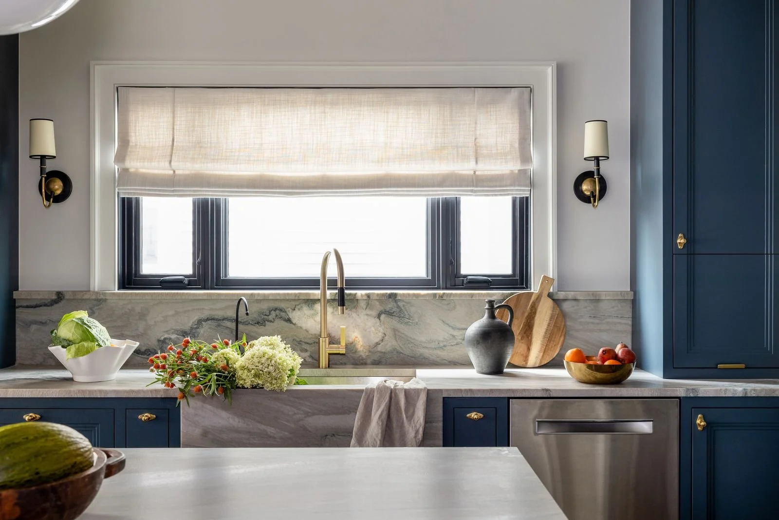 Modern kitchen with navy blue cabinets, a marble countertop, and a window with beige shades. Decor includes a vase, a wooden cutting board, and a bowl of apples.