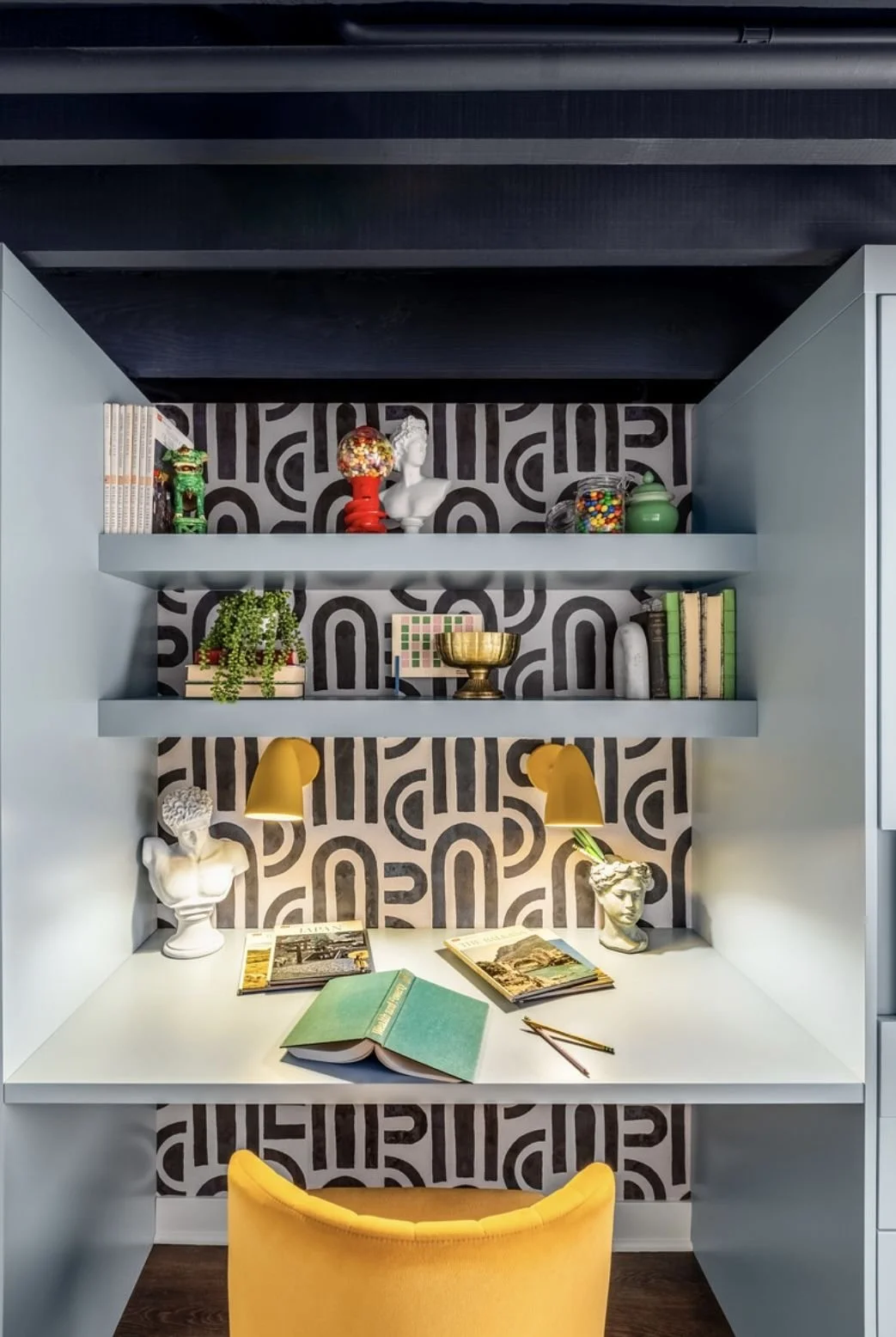A stylish home office with white built-in desk and shelves, black patterned wallpaper, decorative busts, plants, books, art supplies, and yellow accents including a chair and wall sconces.