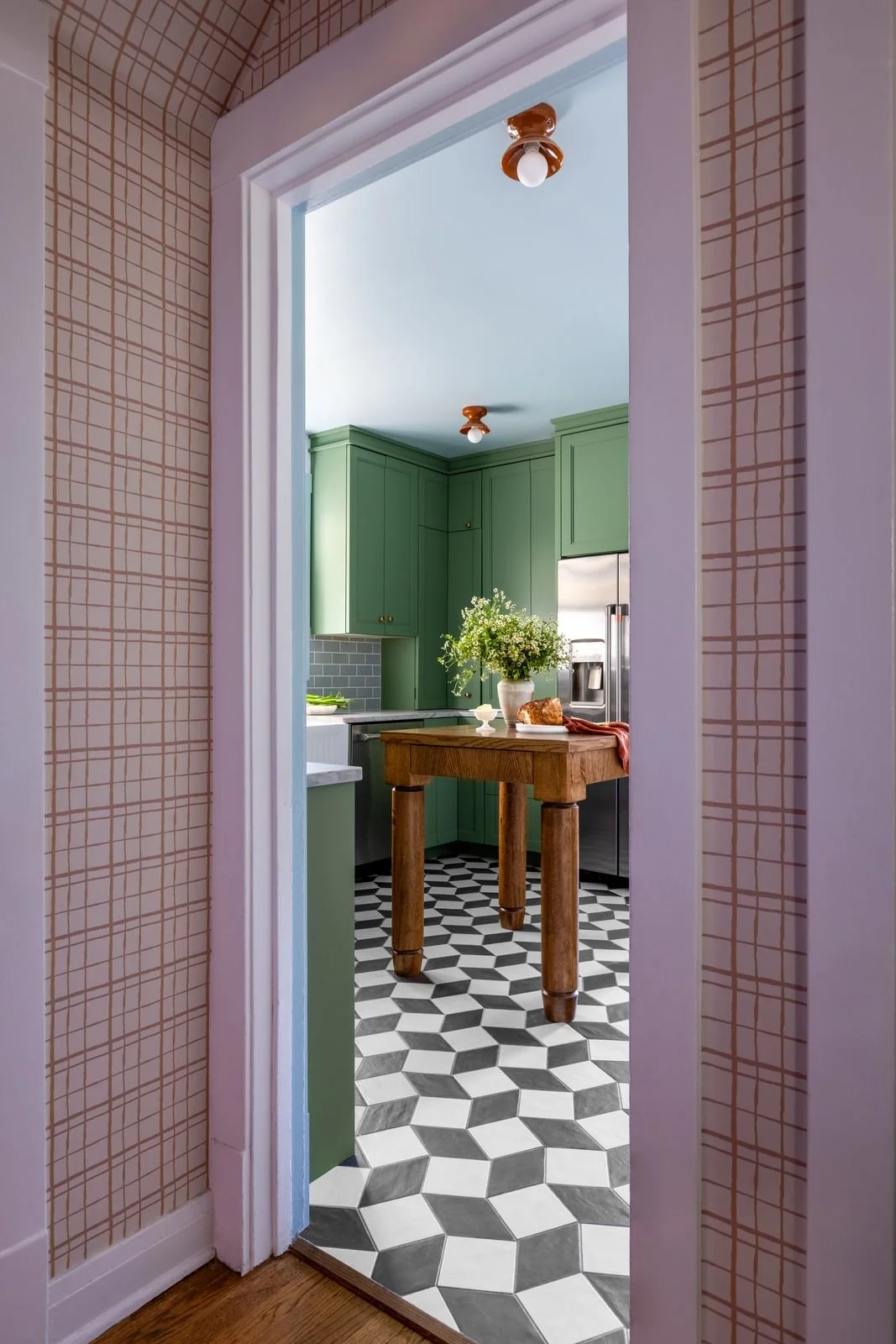 View through a doorway into a kitchen with green cabinets, a wooden table, a vase of flowers, and a patterned tile floor.