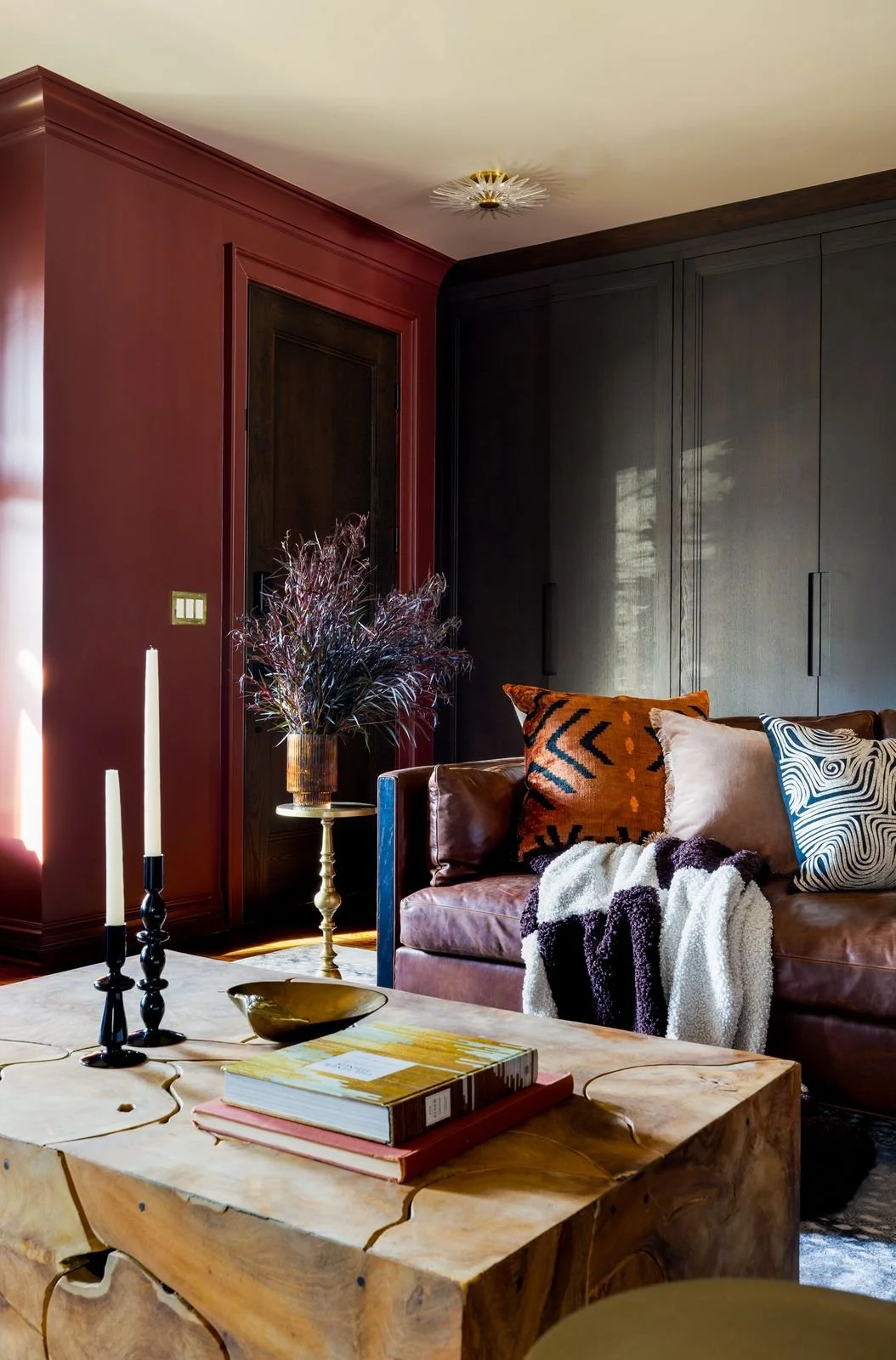 Living room with a red wall, black and brown cabinetry, a leather sofa with assorted pillows and a black and white blanket, a wooden coffee table with books and decorative items, a side table with a dried plant arrangement, and two black candlesticks