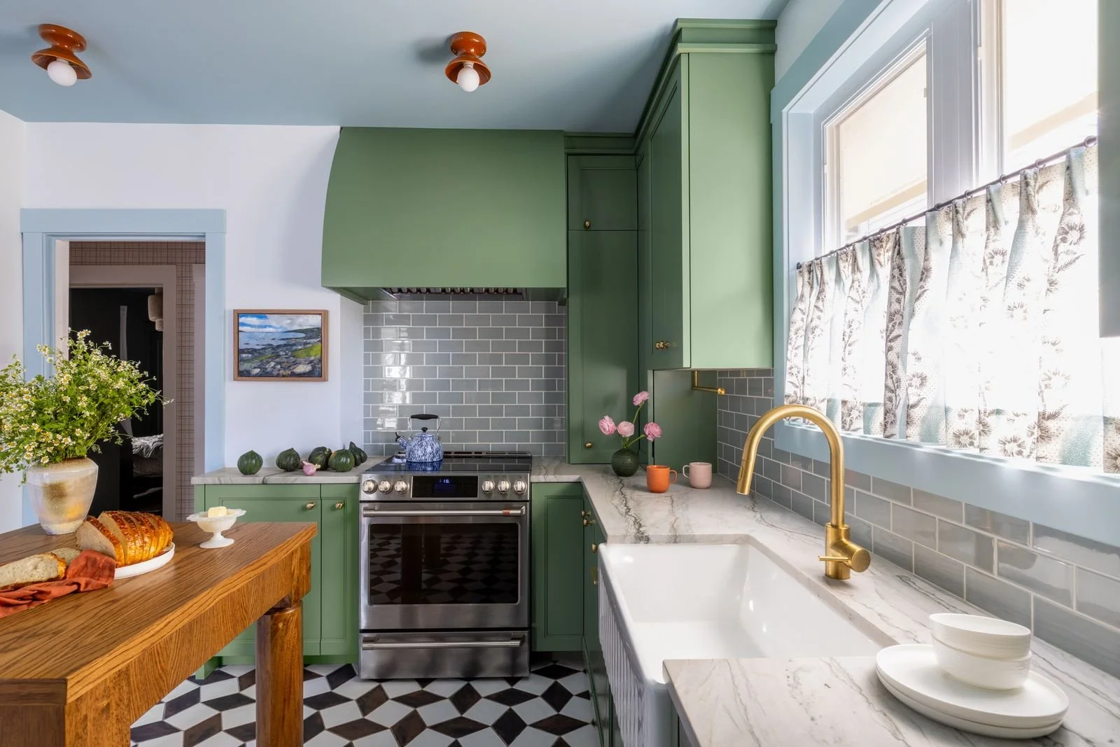 A kitchen with green cabinets, a marble countertop, gray subway tile backsplash, a window with floral curtains, and a white farmhouse sink.