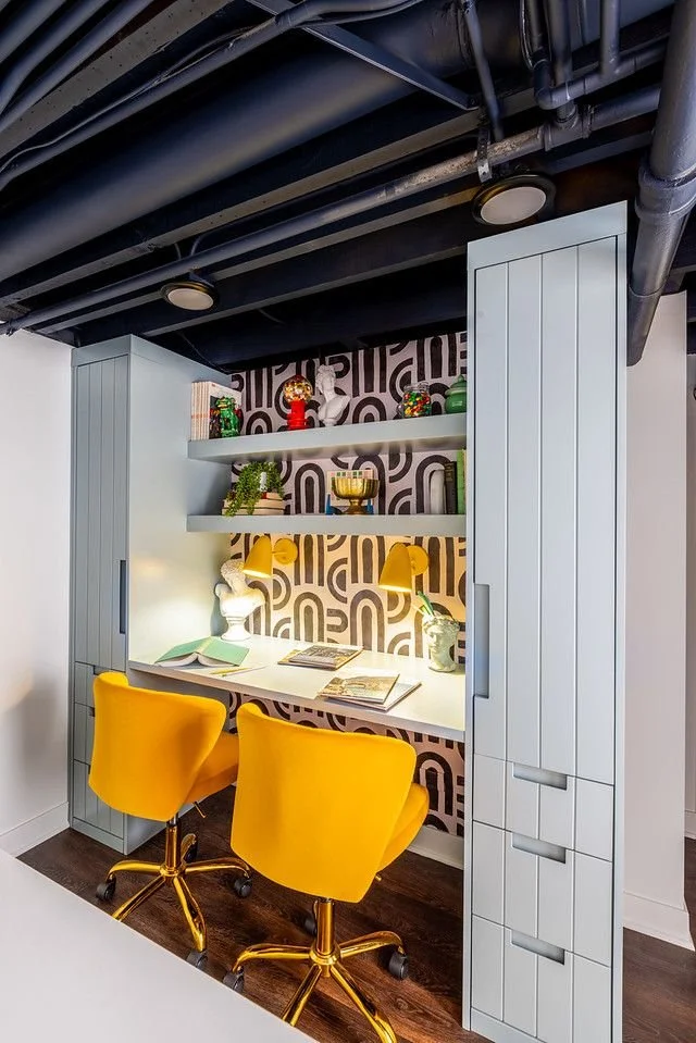 A modern workspace with a built-in desk, two yellow chairs, and open shelves displaying decorative items in a room with dark ceiling pipes and patterned wallpaper.