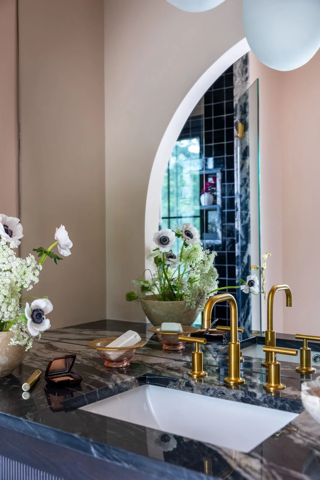 A bathroom countertop with a black marble surface, gold faucets, flowers in vases, and a mirror reflecting a window and part of the room.
