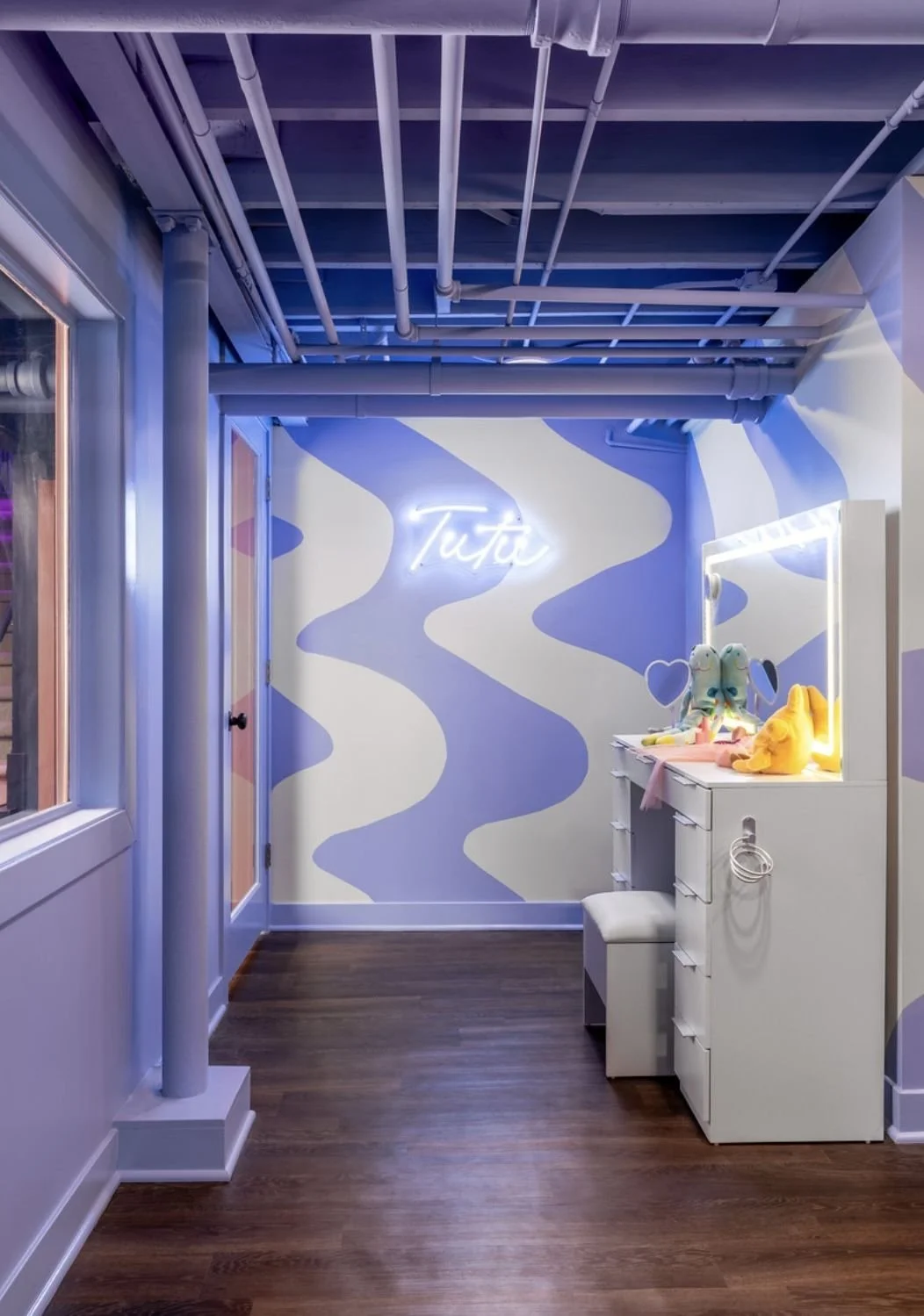 A cozy pastel purple and white corner with a neon sign that says 'Tuta', a mirror with light, stuffed animal toys on a white dresser, and a small stool. The wall has abstract purple and white wave patterns, and the floor is dark wood.
