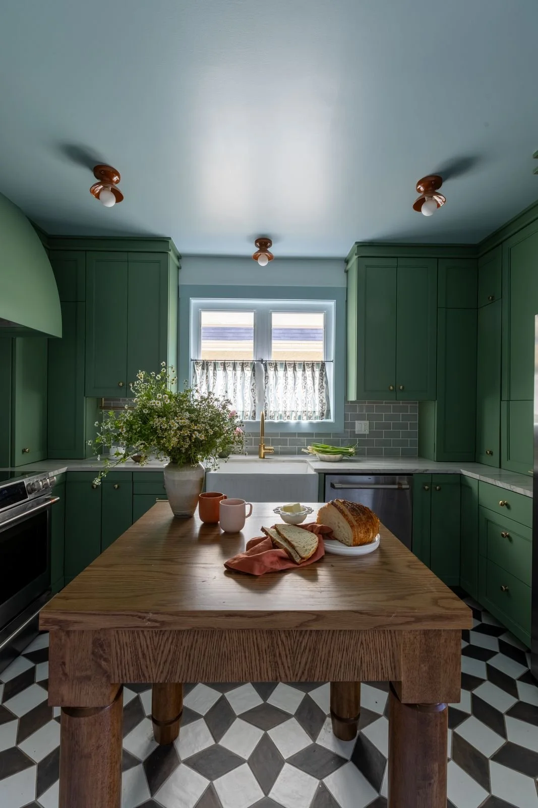 Kitchen with green cabinets, wooden island, checkered black and white floor, window with patterned curtain, flowers, bread, mugs on the island.