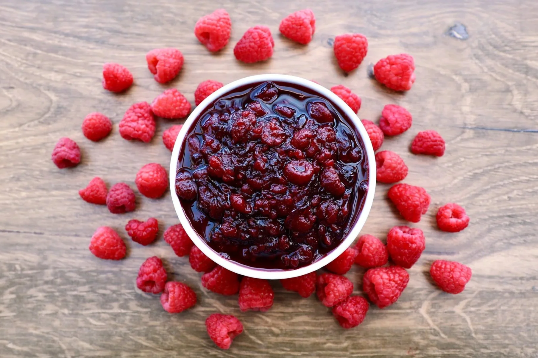 Raspberry inclusions in bowl surrounded by fresh raspberries on a wood surface