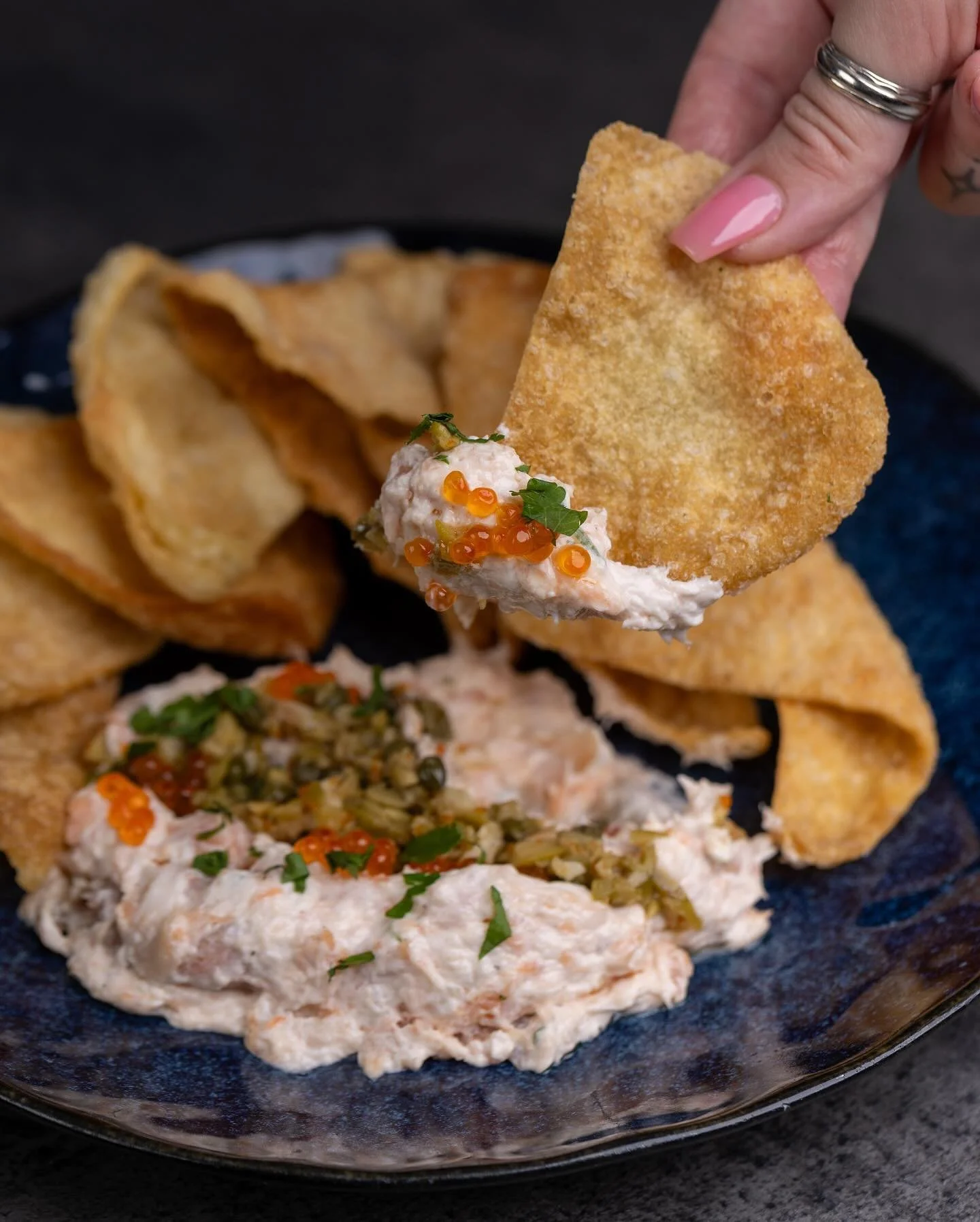 Savor our Smoked Salmon Spread: house-made smoked salmon dip with puttanesca blanco, topped with smoked trout roe, and served with crispy wonton chips. A delightful blend of flavors and textures.