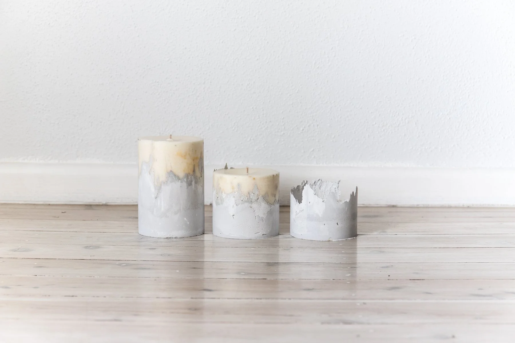 Three cement and wax candles of varying heights on a wooden floor against a white wall.