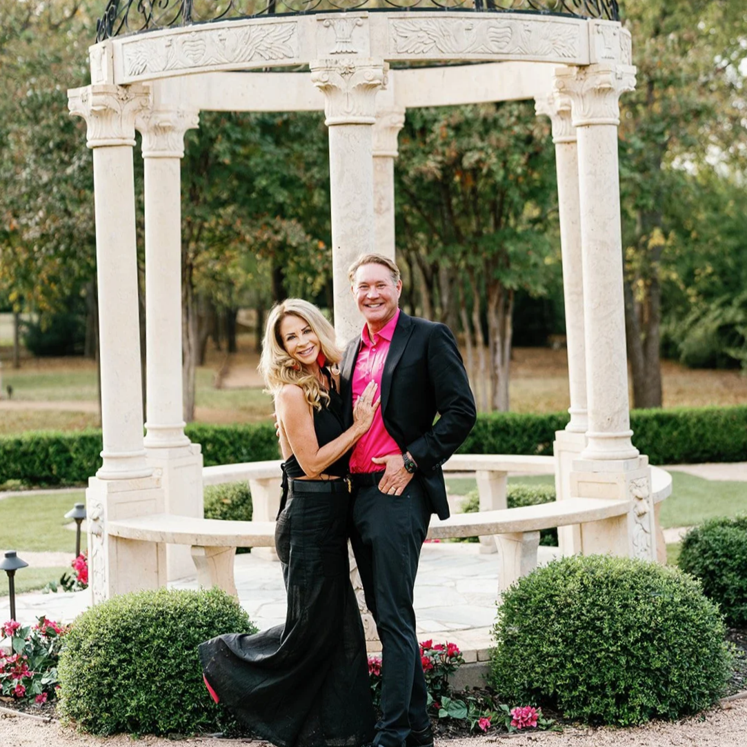 Brian and Becky Banner standing together in front of a decorative stone gazebo in a garden, with trees and bushes in the background.