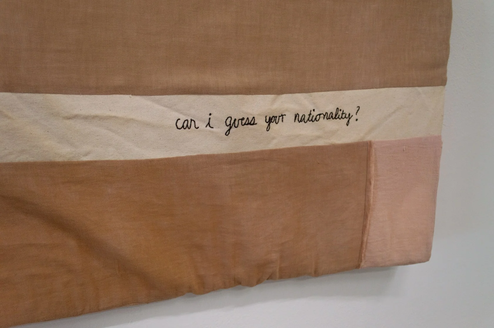 Image: Work on view at Real Tinsel. A beige, color-blocked, fabric painting is embroidered with the words “can i guess your nationality?” Work by Melissa K. Mursch. Photo by Cristina Ossers.