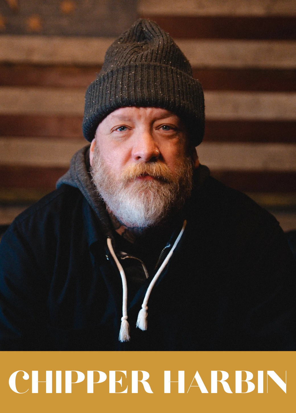 A man with a grey beard and mustache wearing a dark grey knit beanie, a dark hoodie, and a black jacket. He has a nose piercing and is looking directly at the camera with a serious expression. The background shows wooden planks. Text at the bottom reads "CHIPPER HARBIN."