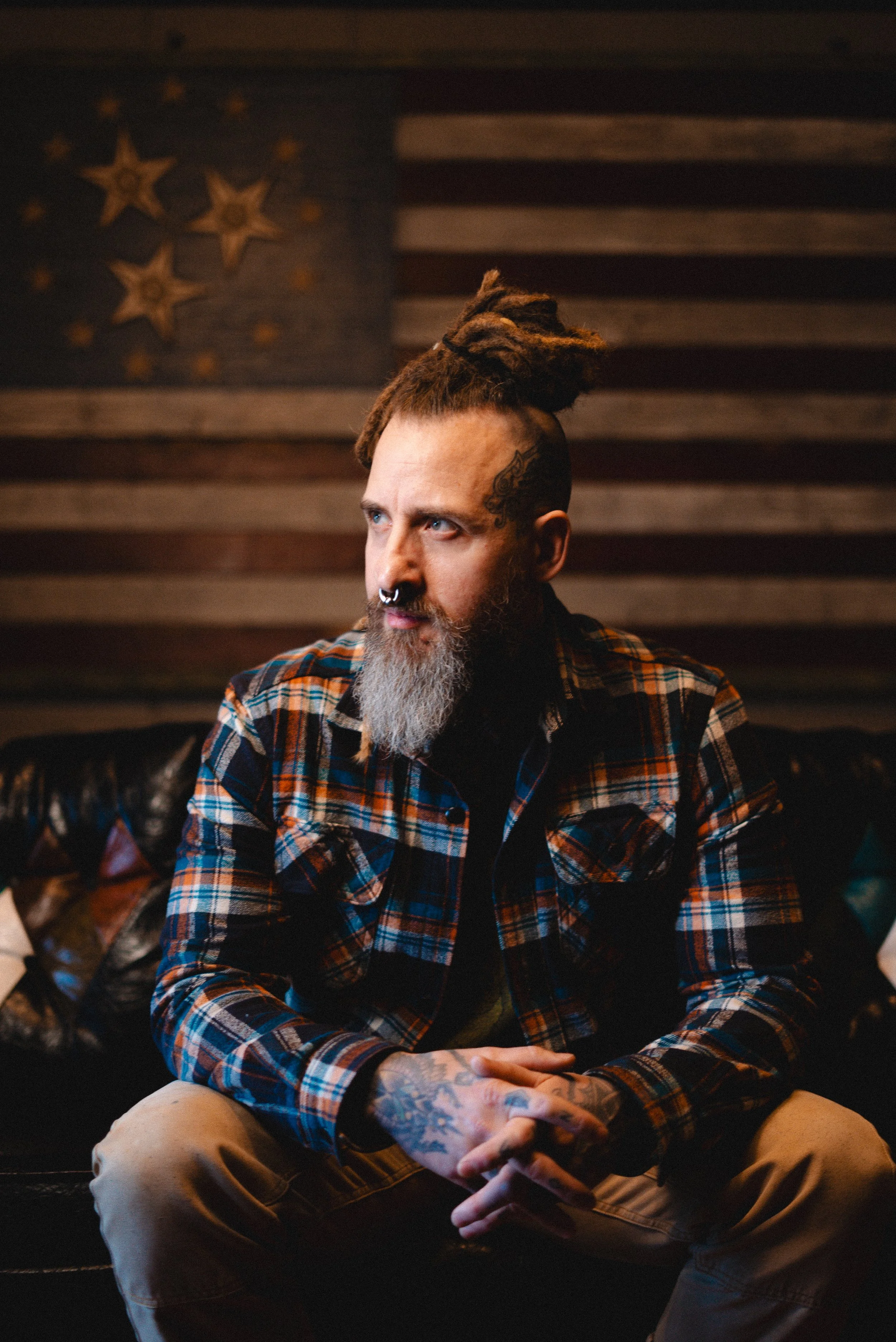 A man with a beard and dreadlocks hairstyle with shaved sides, sitting on a black leather couch, wearing a plaid shirt, with a tattooed hand, in front of an American flag wall.