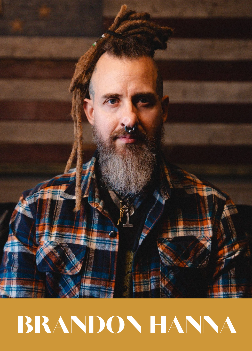 A man with dreadlocks, a gray beard, and a septum piercing, wearing a plaid shirt, sitting against a wooden background.