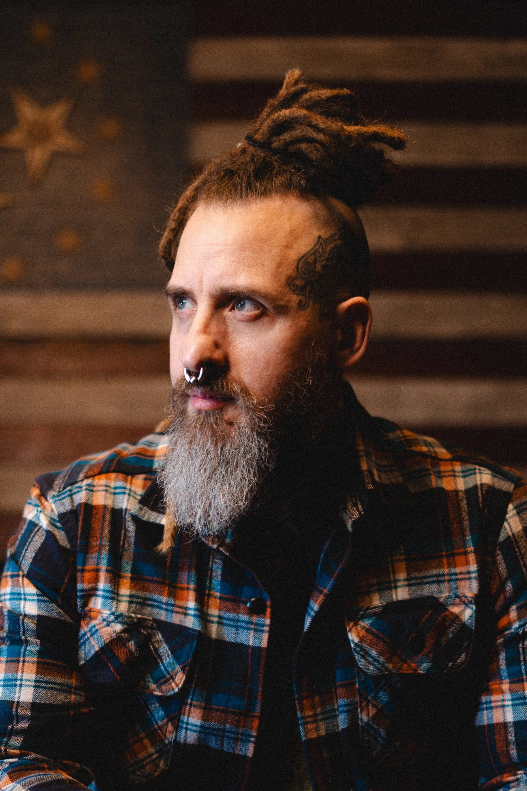 A man with dreadlocks, a beard, and a septum piercing, wearing a plaid shirt, looking to the side with a serious expression. The background includes a wooden wall with star-shaped decorations.