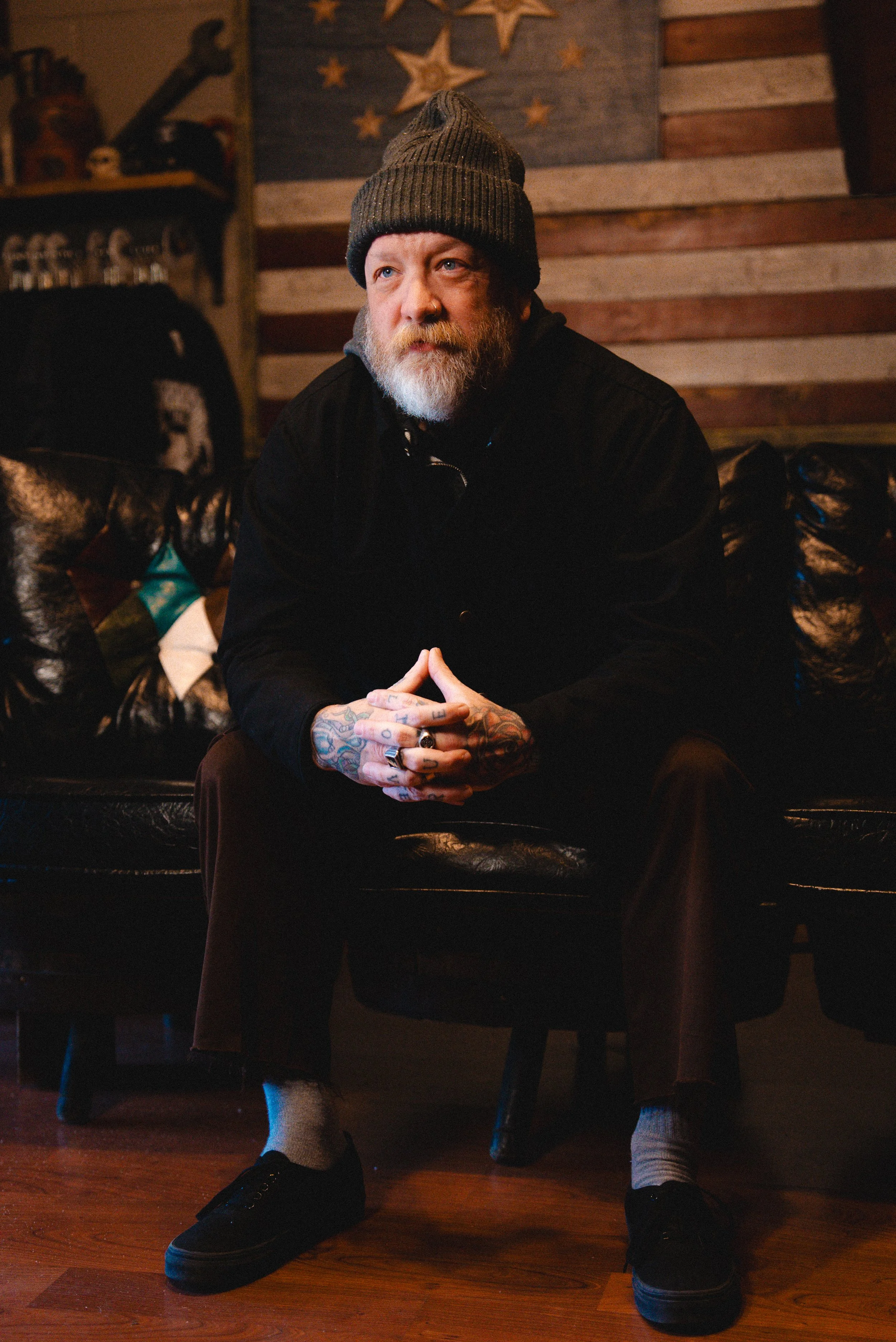 A bearded man wearing a gray beanie, black jacket, and gray socks with black shoes, sitting on a black leather couch with his hands clasped, in a room with a rustic wooden wall and decorative star-shaped ornaments.