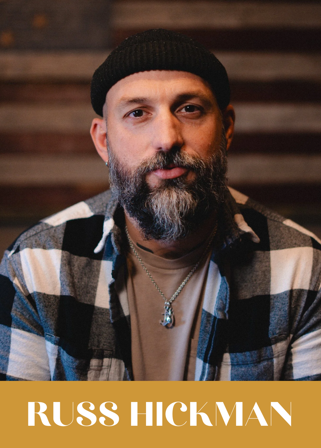 A man with a beard and mustache wearing a black beanie, a checkered shirt over a t-shirt, and a silver chain necklace with a pendant. The background has wooden slats. The text overlay at the bottom reads 'RUSS HICKMAN' in white capital letters on a gold background.