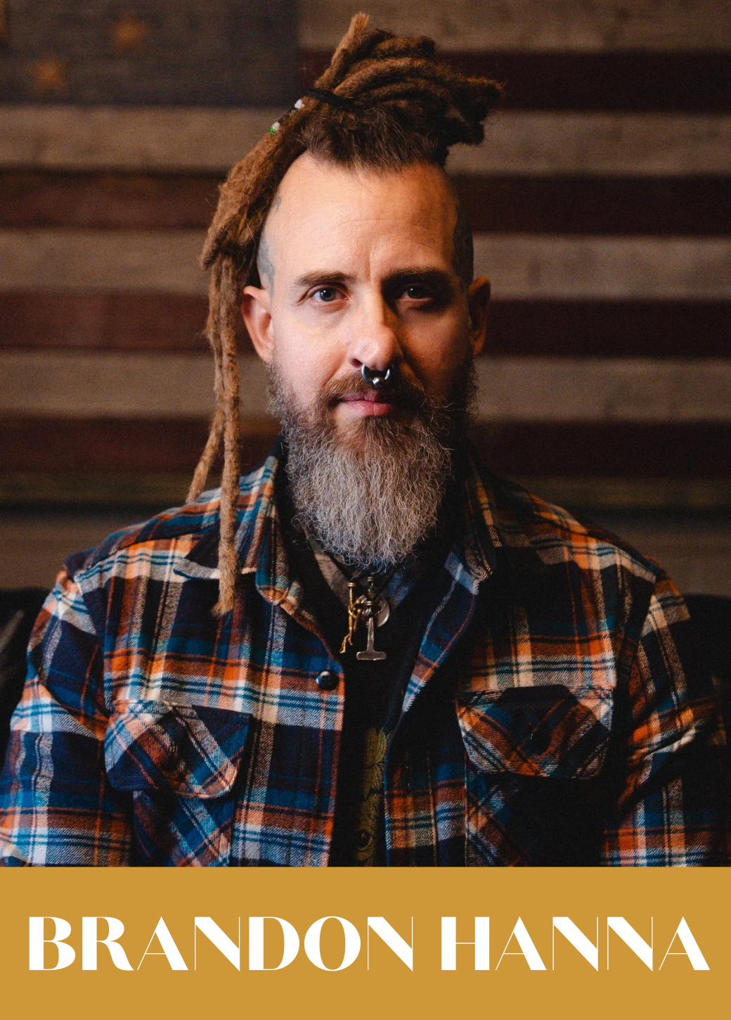 A man with dreadlocks, a beard, and a septum piercing wearing a plaid shirt and a large cross necklace, sitting in front of a wooden background.