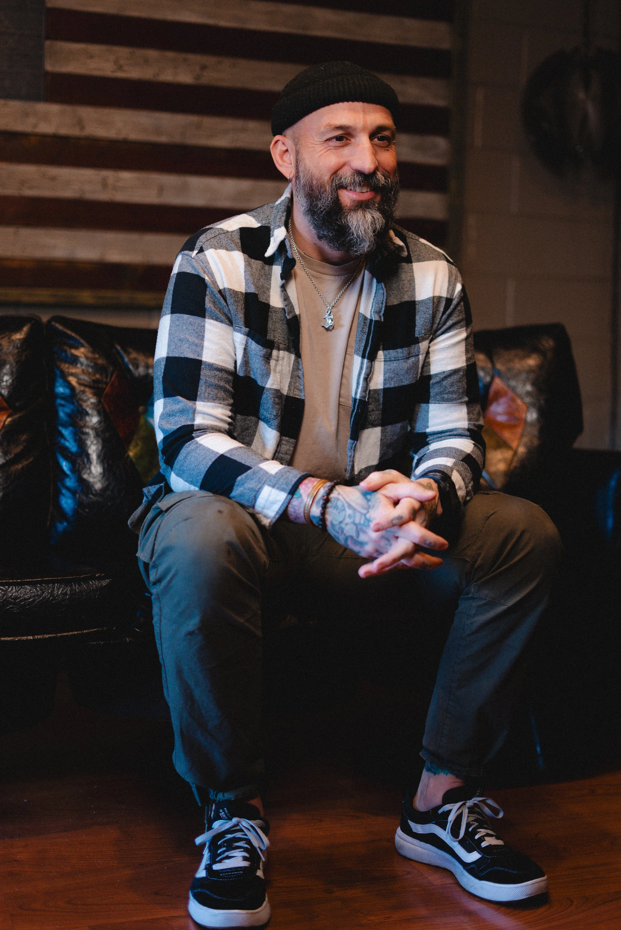 A man with a beard and tattoos, wearing a black beanie, checkered shirt, beige t-shirt, and black pants, sitting on a black leather couch in a room with wooden wall decor.