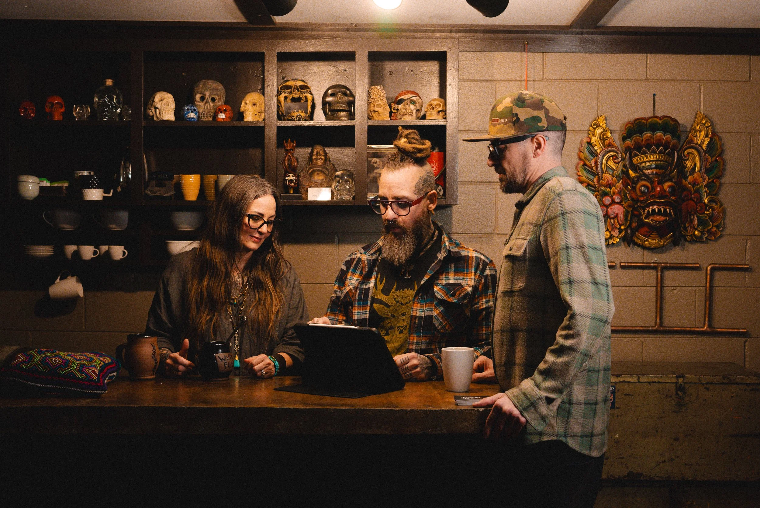 Three people gathered around a counter, looking at a tablet in a cozy, eclectic room with shelves of various decorative skulls and masks on the wall behind them.