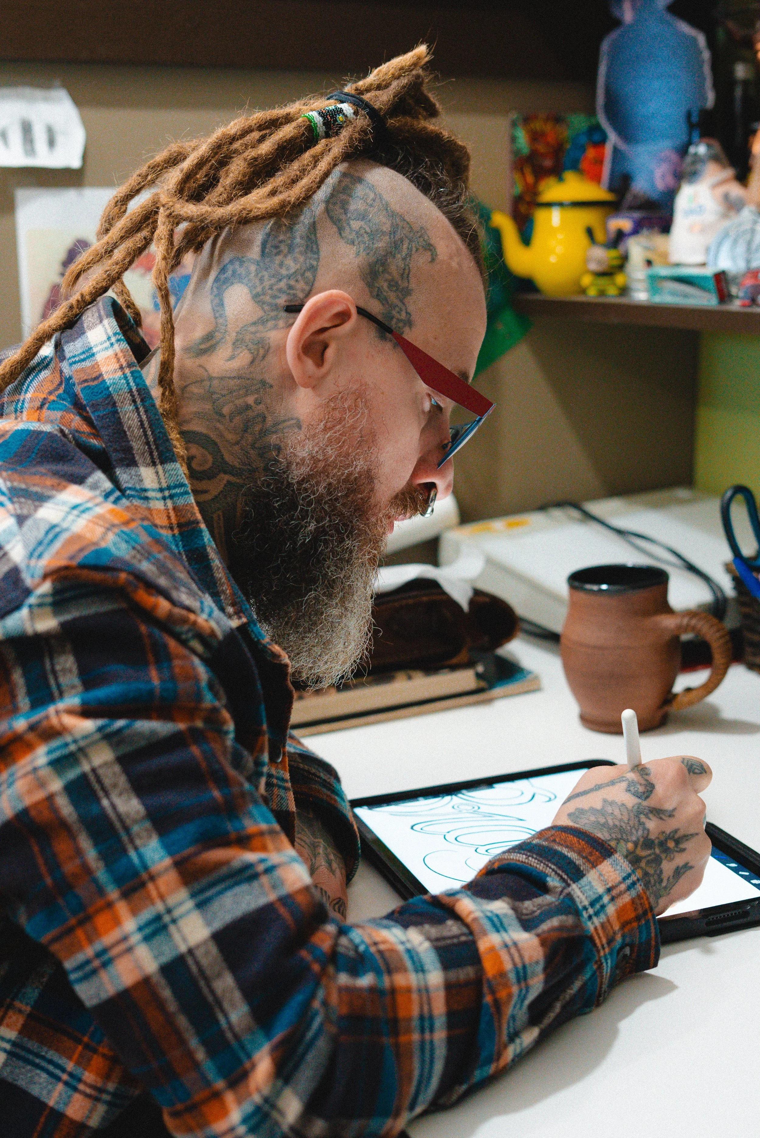 A tattooed man with dreadlocks, glasses, and a plaid shirt drawing on a digital tablet with a stylus.