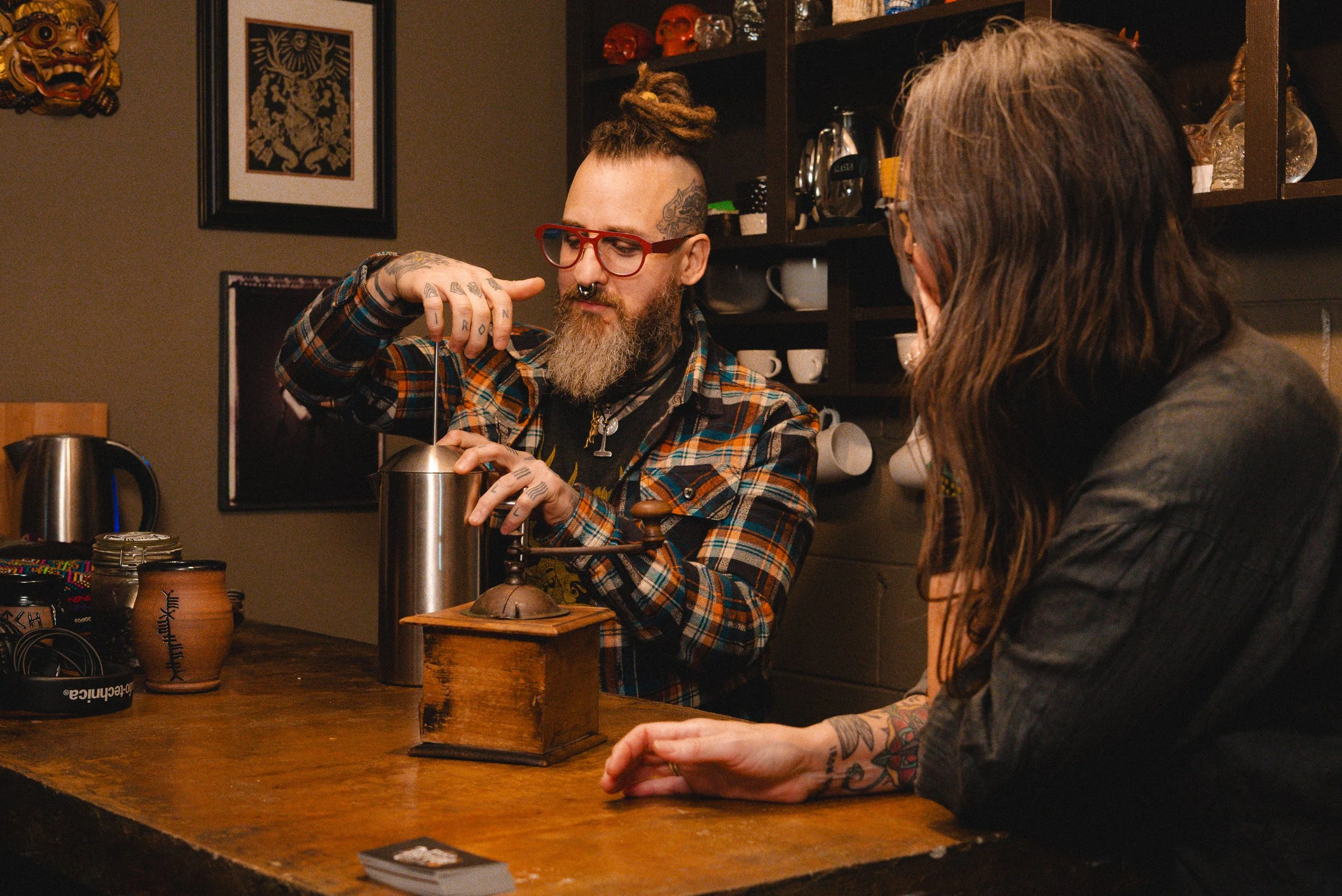 A tattooed man with glasses and dreadlocks operating a vintage coffee grinder at a wooden table while looking at a woman with tattoos on her arm, who is sitting across from him. The background has shelves with cups, glasses, and decorative skull scul