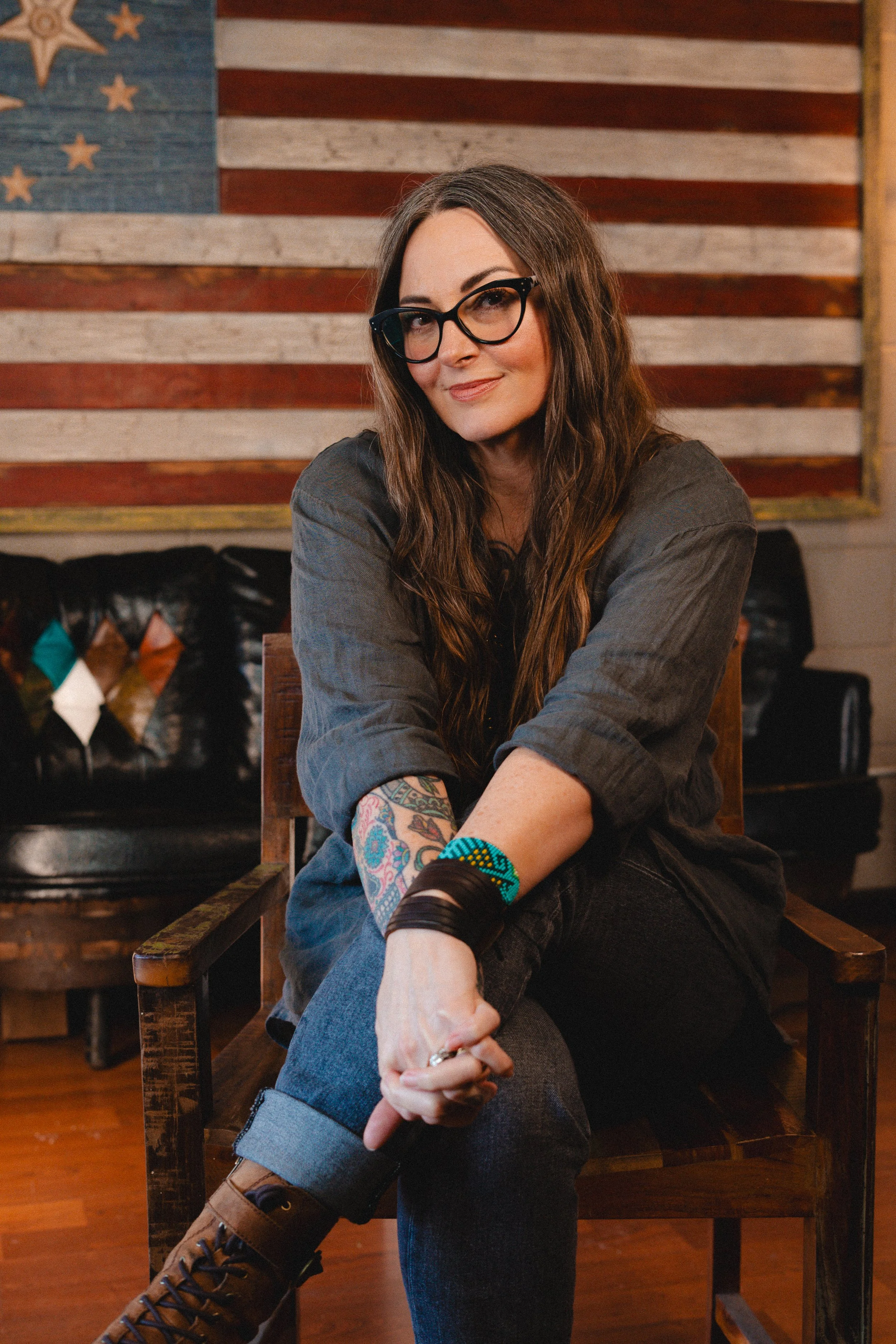 A woman with long wavy brown hair and glasses sitting on a wooden chair inside a rustic room, with a patriotic American flag decoration on the wall behind her.