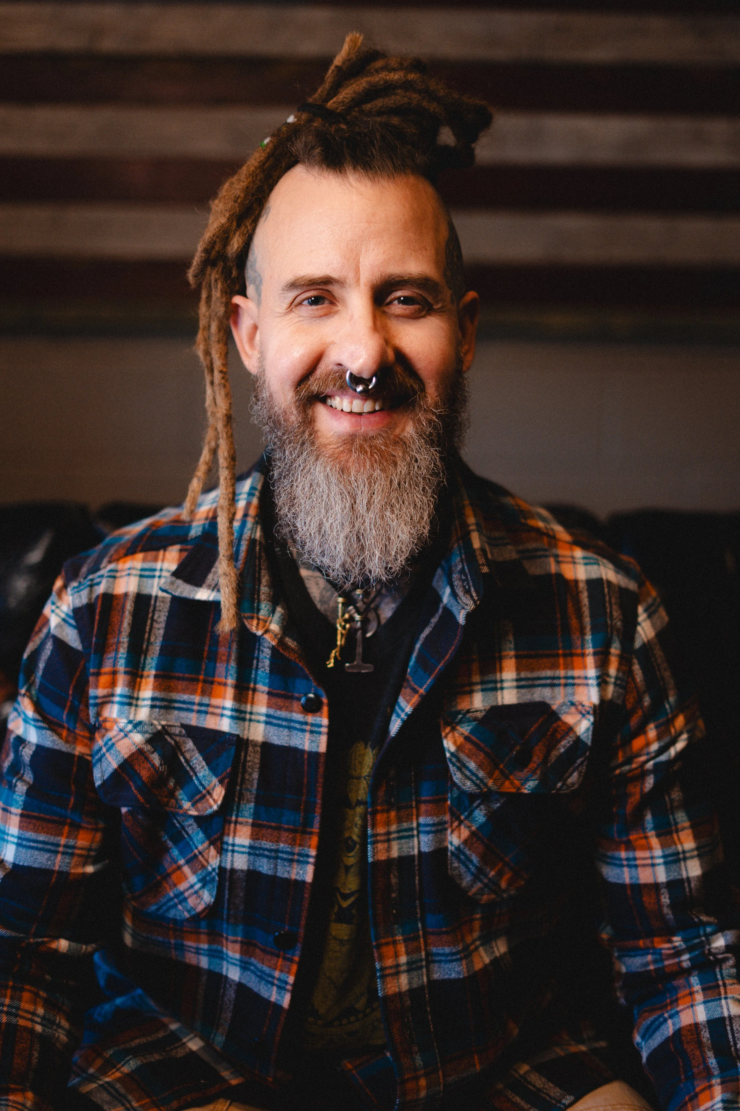 Man with dreadlocks, beard, tattoo, septum piercing, colorful plaid shirt, and necklace, smiling indoors.