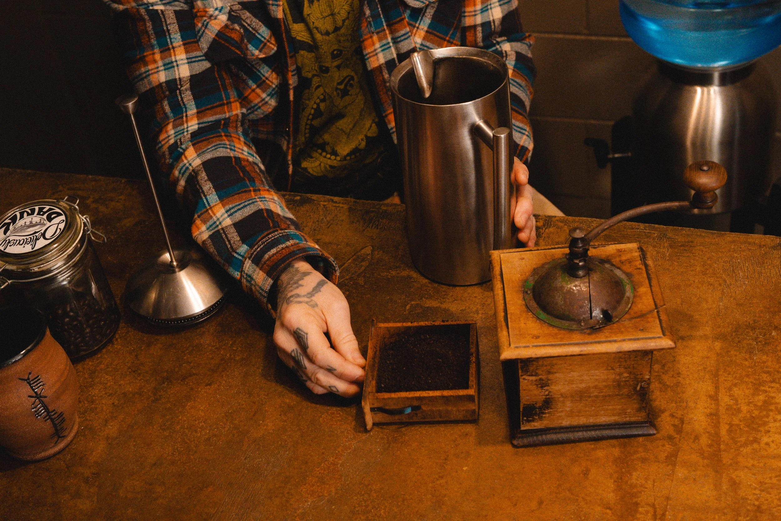 Person with tattoos in a plaid shirt holding a metal container, next to a wooden coffee grinder on a rustic wooden counter. Coffee beans and jars are also visible.