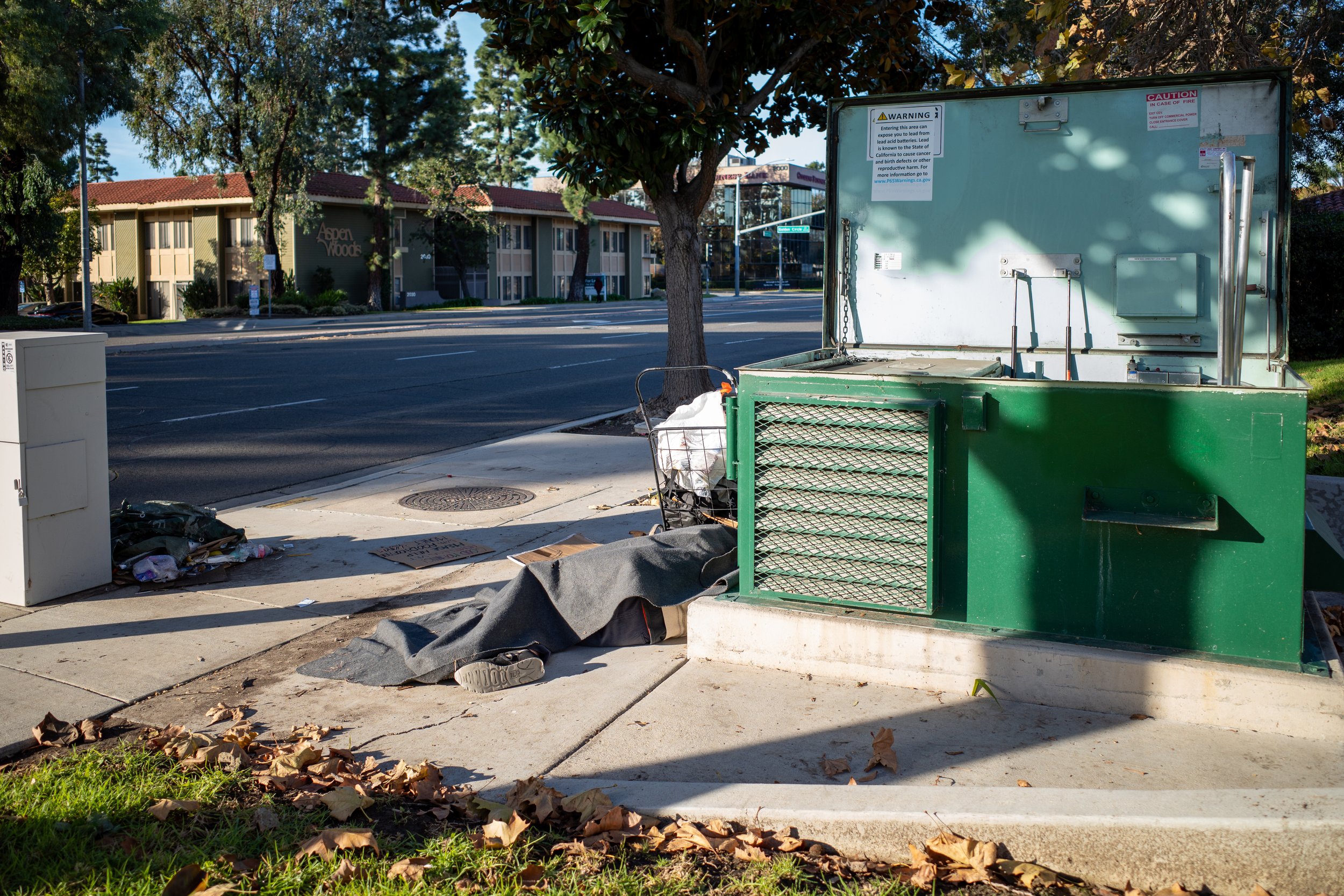 Photo of a man sleeping on the sidewalk next to a green electrical transformer with a shopping cart parked near his head.