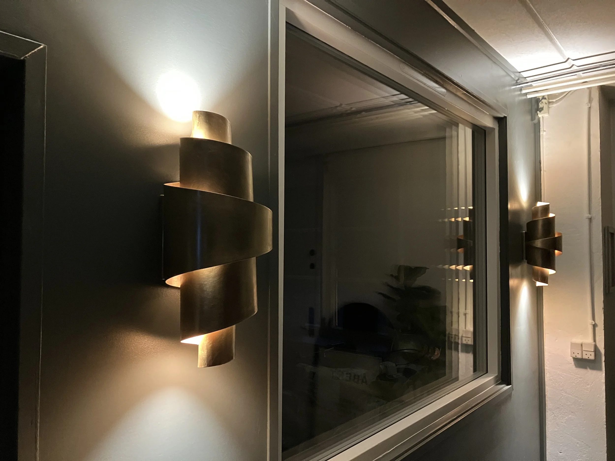 a pair of softly glowing brass sconces inspired by the roll-shape of a croissant flank a large window in a wall in a bakery