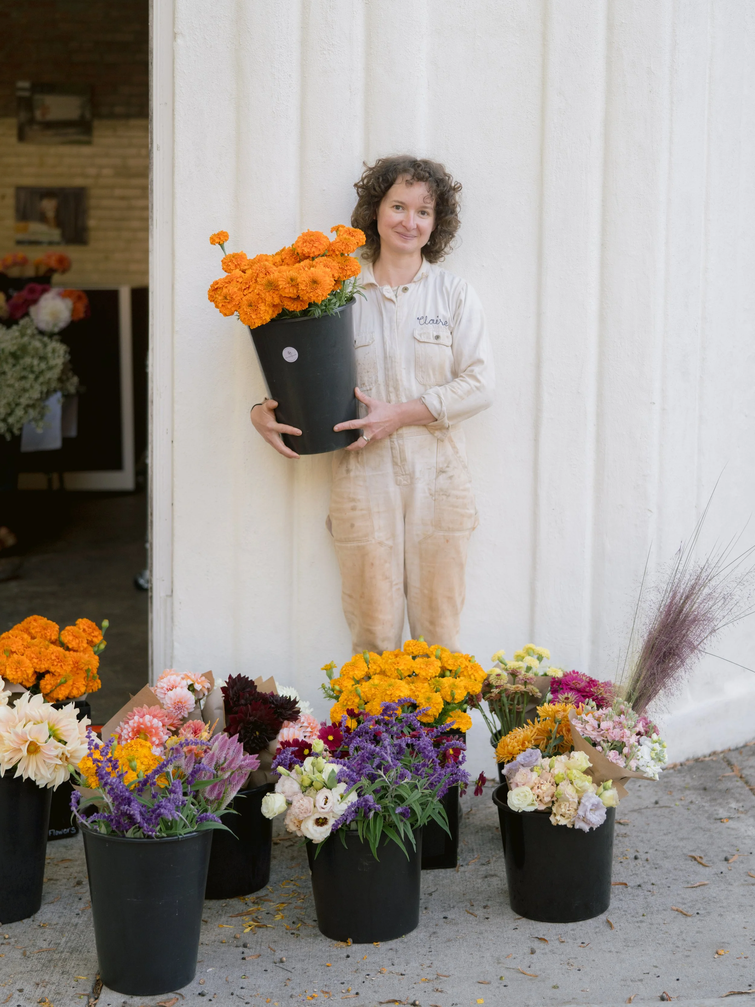 A woman with curly hair, wearing a cream-colored jumpsuit with her name 'Claire' embroidered on the chest, holding an orange marigold flower pot, standing behind a display of various colorful flowers in black pots.