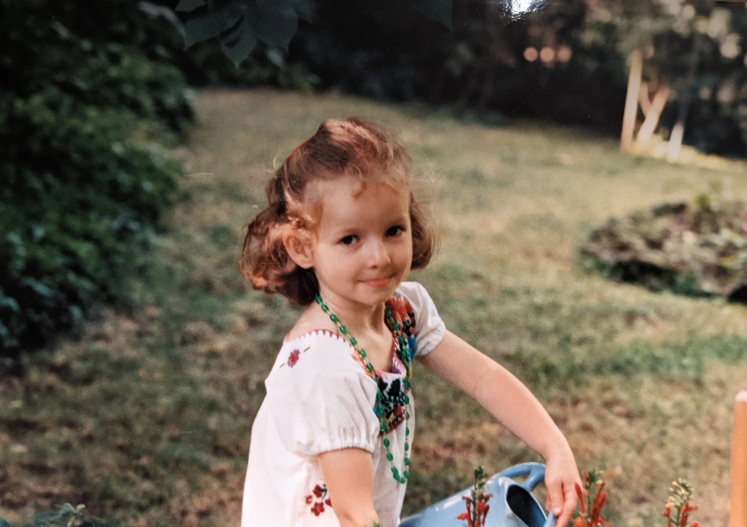 A young girl with curly red hair wearing a white dress with embroidered flowers, standing outdoors during dusk or evening, surrounded by greenery and trees, with a smile on her face.