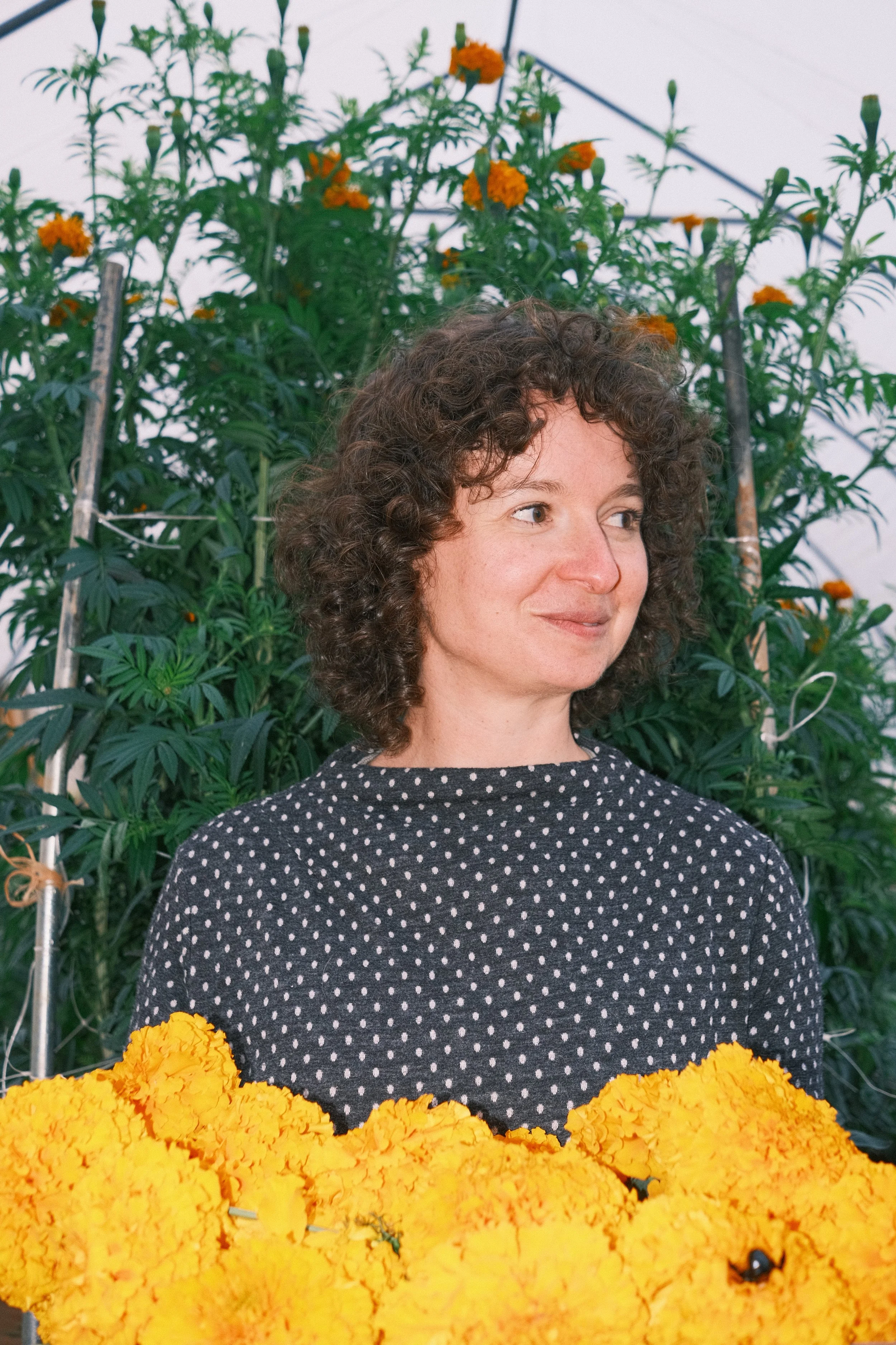 A woman with curly brown hair and light skin smiling while standing in front of green foliage. She is wearing a black dress with white polka dots. In the foreground, there are bright yellow-orange marigold flowers.