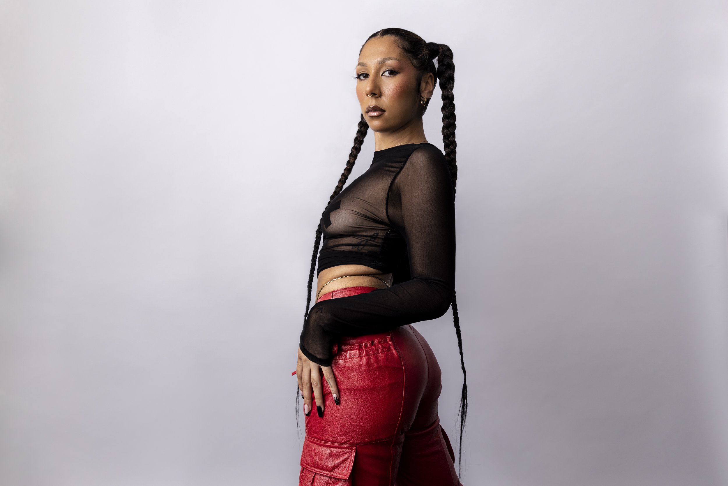 A studio photograph of the singer Miss Kaninna wearing her signature braided pigtails, wearing a long-sleeved meshed crop top and red leather trousers.