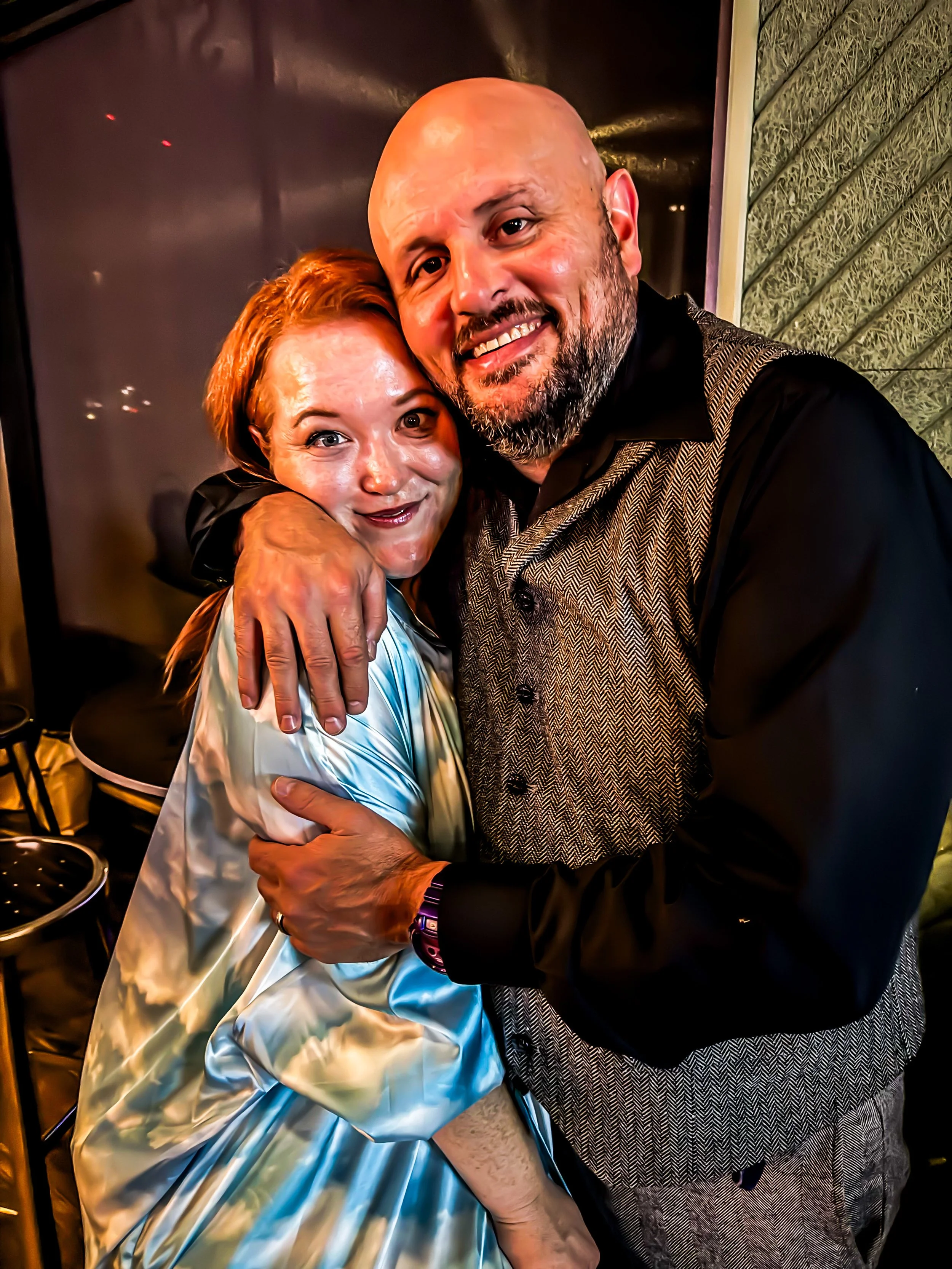 A photo of Jesse James Ziegler hugging his wife, Amanda McHenry. Jesse is wearing a grey tweed suit vest with a black shirt, and Amanda is wearing a blue silk dress.