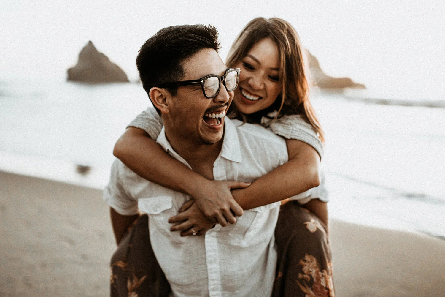 A joyful couple at the beach, with the woman hugging the man from behind, both smiling and laughing. Photographed by Will Khoury.