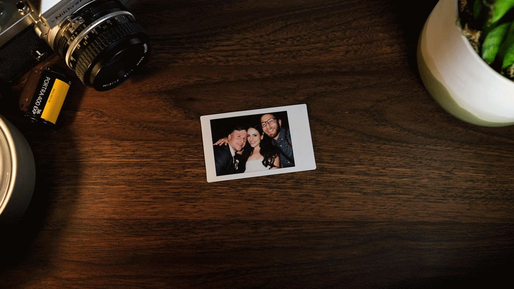 A small printed photograph of three people, placed on a wooden surface surrounded by a camera, film roll, and a potted plant.