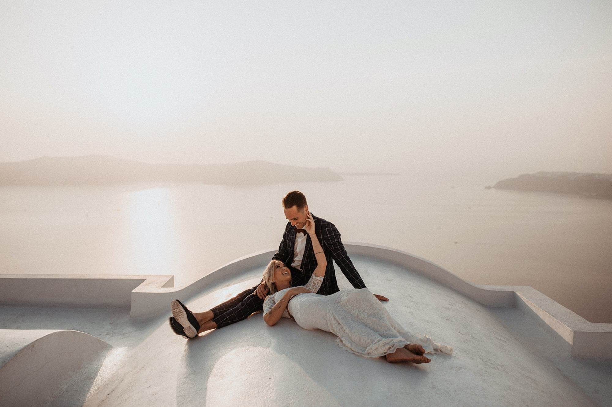 A couple is on a white rooftop, with the woman lying down and the man sitting beside her, smiling and touching her face, overlooking a body of water and distant islands at sunset.