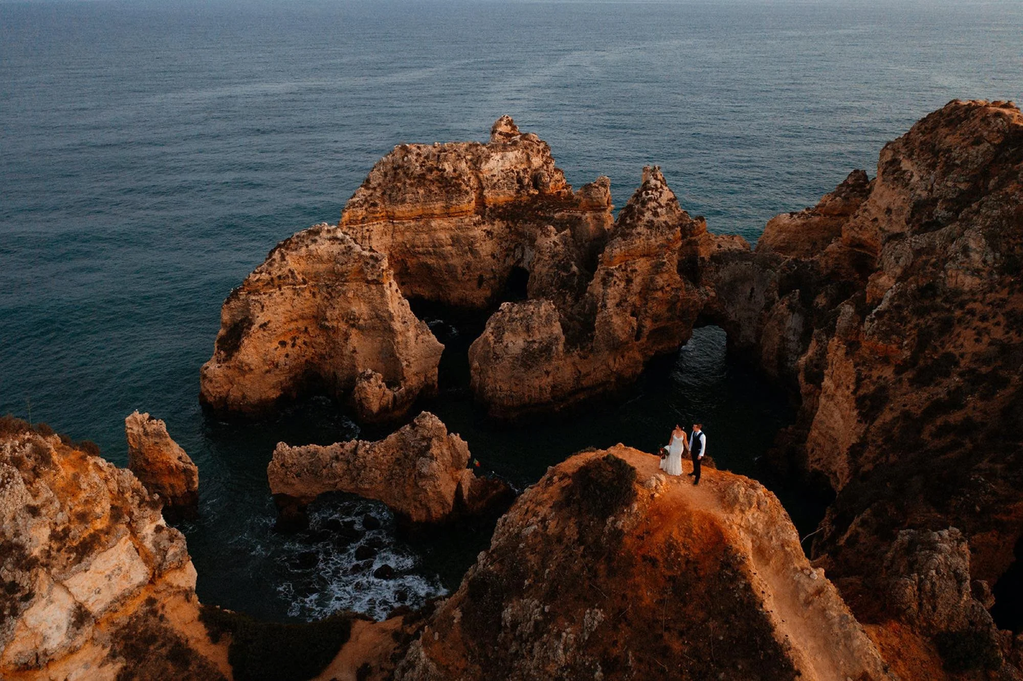 A couple standing on a rocky cliff overlooking the ocean, with large rock formations and arches nearby, during sunset.