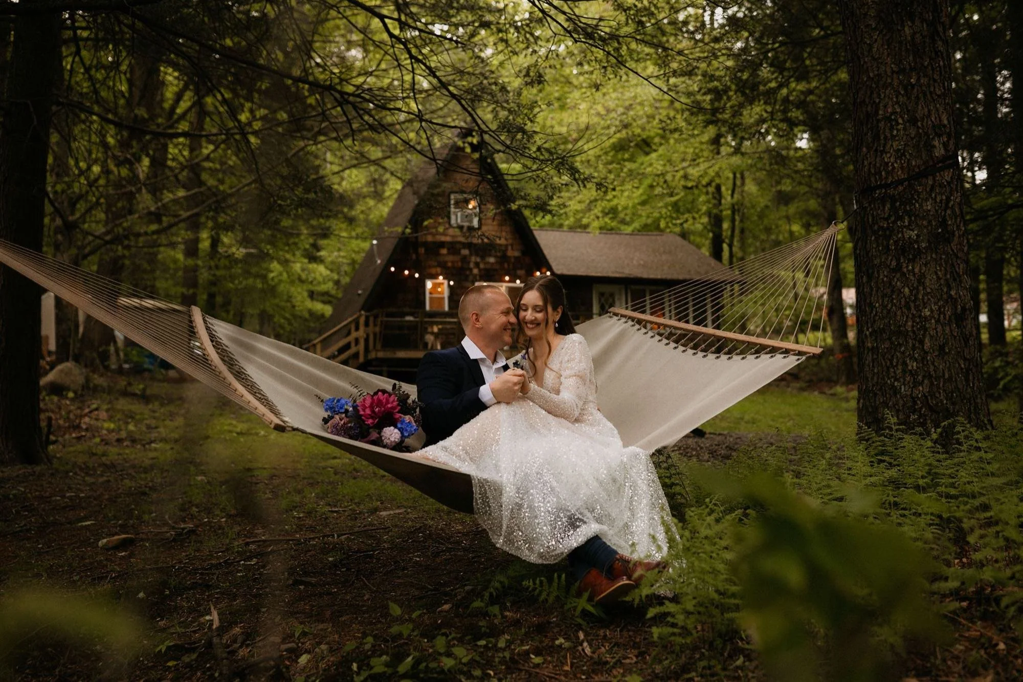 A newlywed couple sitting together on a hammock in a forested area with a rustic cabin in the background, during the evening.