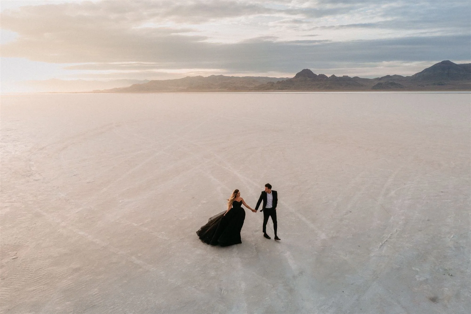 A couple dressed in formal attire holding hands and walking across a vast, flat white desert landscape with mountains in the background and a cloudy sky.