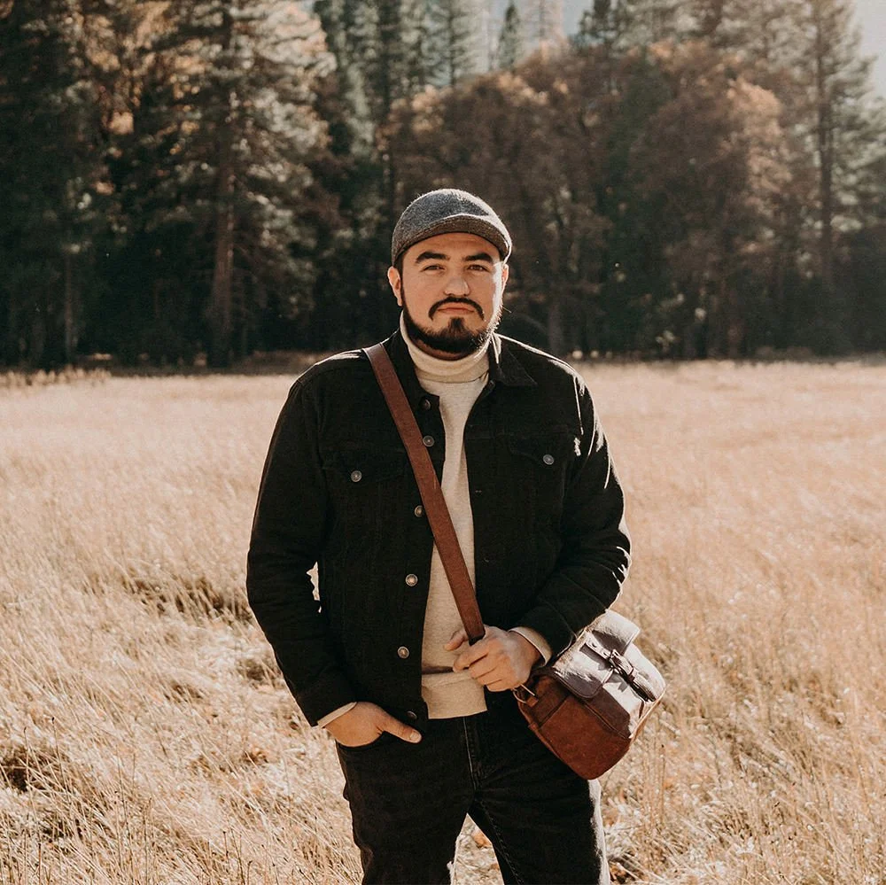 A man with a beard and mustache standing outdoors in a field of tall, dry grass with trees in the background, wearing a gray beanie, black jacket, and carrying a brown bag.