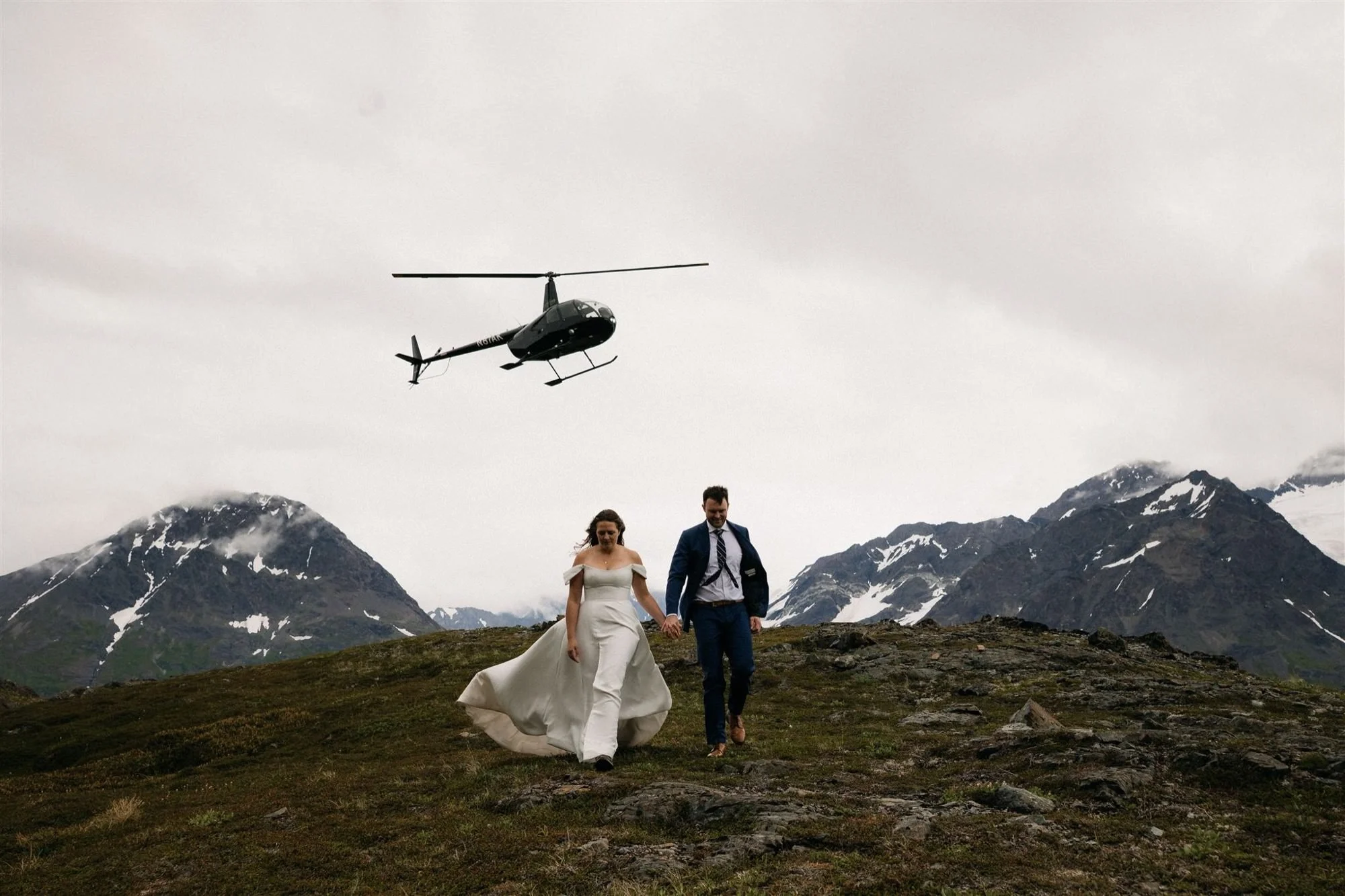A bride and groom walking hand in hand on a grassy mountain landscape with snow-capped peaks in the background, a helicopter flying overhead on a cloudy day.