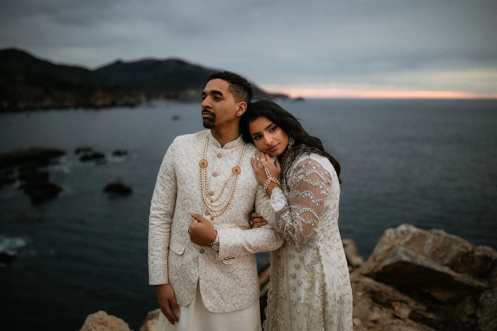 A couple in elegant traditional Indian attire stands near a rocky shoreline at sunset, with water and distant hills in the background. The man wears a cream-colored embroidered sherwani with layered pearl necklaces, and the woman wears an intricate w