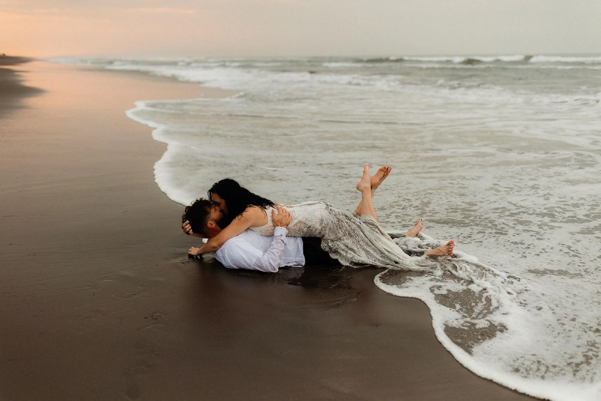A couple lying on the beach near the water, embracing and kissing at sunset.